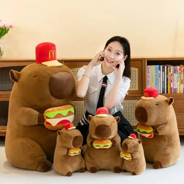 A group of capybara plush toys in various sizes, wearing red hats and holding plush hamburgers, with a person sitting behind them.