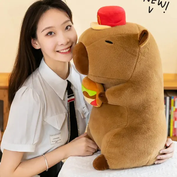 A side profile of a large capybara plush toy wearing a red hat and holding a plush hamburger, posed with a smiling person.