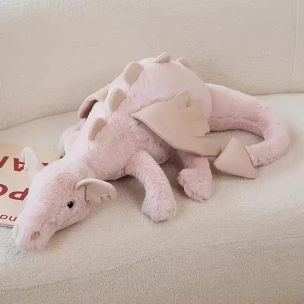 A pastel pink dragon stuffed animal with soft fabric wings and plush spikes, lying comfortably on a couch.