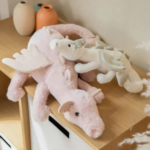 A pink plush dragon stuffed animal with a smaller white dragon plush resting on top, displayed on a wooden shelf.